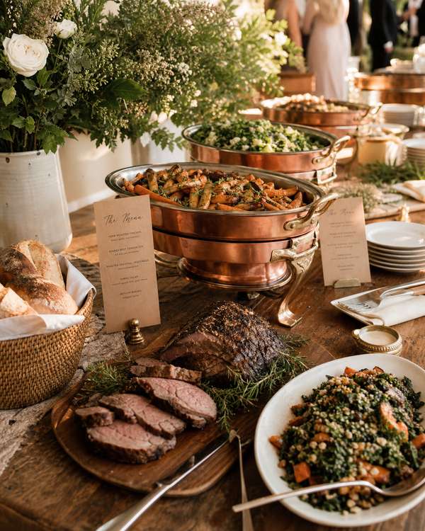 A family-style buffet display with carved meats, roasted vegetables, and bread baskets on a long oak table